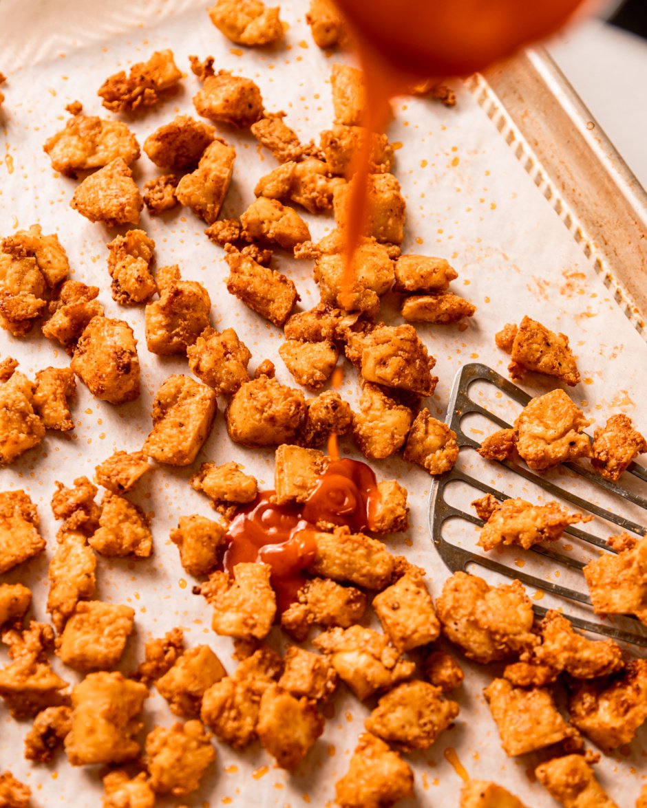An overhead shot shows Buffalo-style hot sauce being poured on top of some crispy little bits of baked tofu.