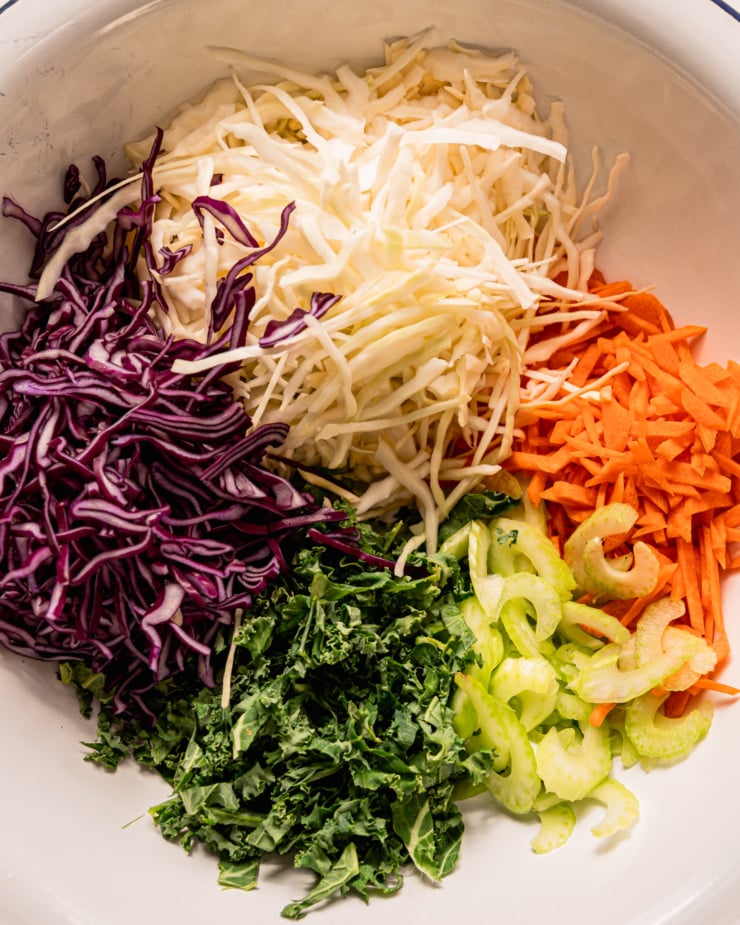 An overhead shot shows the following in a bowl: shredded red and green cabbage, shredded carrots, sliced celery, and sliced kale.