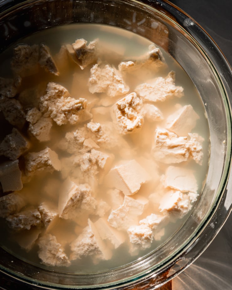 An overhead shot shows pieces of tofu soaking in water in direct, harsh light.