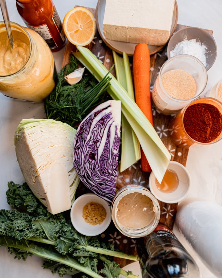 An overhead shot shows ingredients for a vegan ranch slaw with crunchy baked Buffalo tofu: arrowroot starch, garlic powder, paprika, onion powder, carrot, celery, cabbage, maple syrup, cashew butter, avocado oil, apple cider vinegar, nutritional yeast, kale, dill, chives, Dijon mustard, Buffalo-style hot sauce, lemon, and firm tofu.