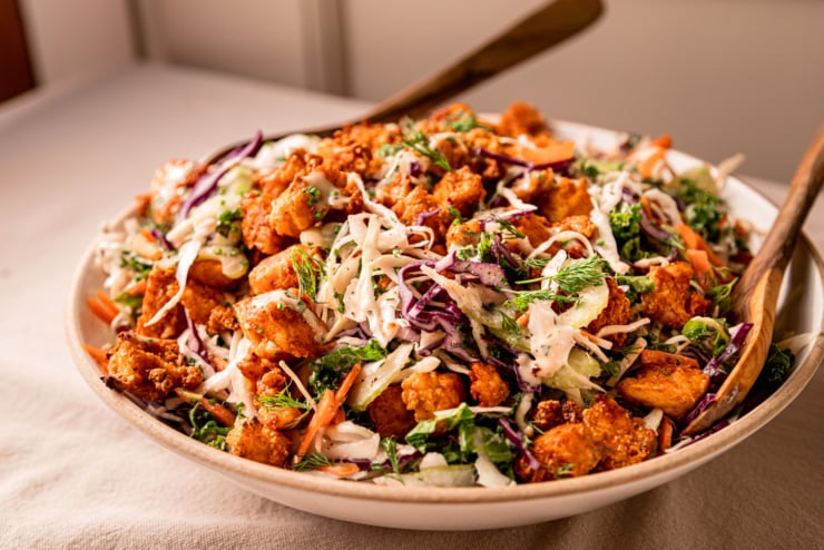 A head-on shot shows a wide serving bowl filled with a colourful vegan ranch slaw. The slaw is topped with crunchy little bits of Buffalo sauce-coated tofu. Wooden tongs are sticking out of the bowl.