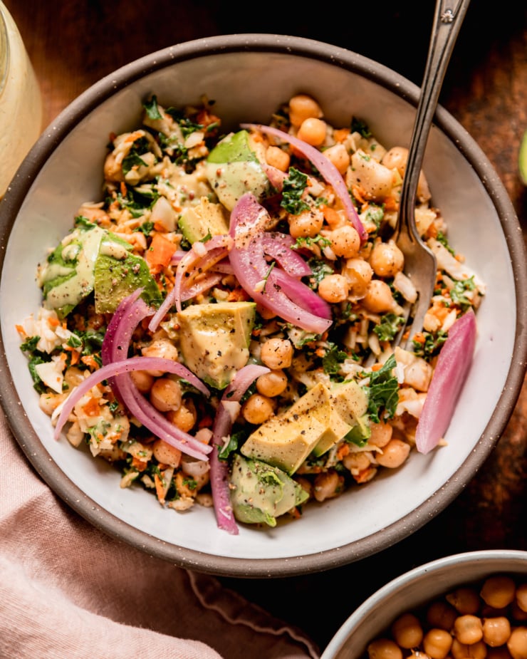 An overhead shot shows an individual serving of a chopped kale cabbage salad with chickpeas, avocado, pickled red onion, and sweet mustard dressing. A fork is sticking out of the bowl.