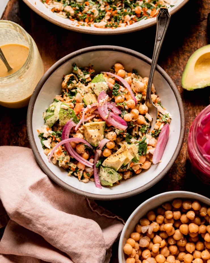 An overhead shot shows an individual serving of a chopped kale cabbage salad with chickpeas, avocado, pickled red onion, and sweet mustard dressing. A fork is sticking out of the bowl. Individual components of the salad can be seen nearby.