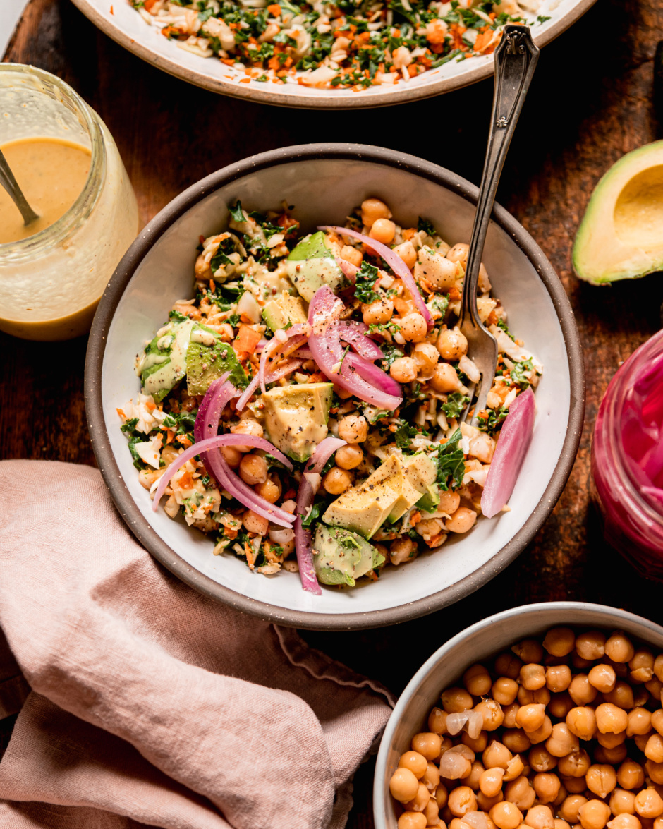 An overhead shot shows an individual serving of a chopped kale cabbage salad with chickpeas, avocado, pickled red onion, and sweet mustard dressing. A fork is sticking out of the bowl. Individual components of the salad can be seen nearby.