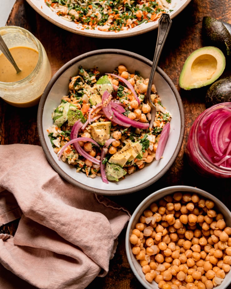 An overhead shot shows an individual serving of a chopped kale cabbage salad with chickpeas, avocado, pickled red onion, and sweet mustard dressing. A fork is sticking out of the bowl. Salad components are nearby and a napkin is bunched up around the serving bowl.