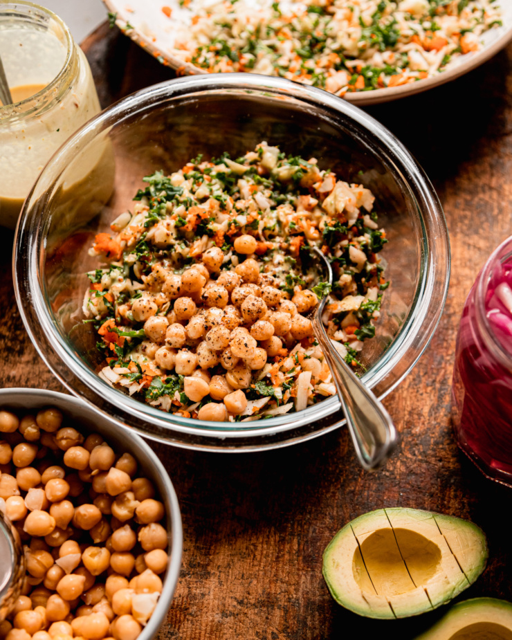 A 3/4 angle shot shows a mixture of chopped kale, cabbage, and carrots being mixed in a bowl with chickpeas and a sweet mustard dressing. A halved, diced avocado is nearby as well as a jar of dressing.