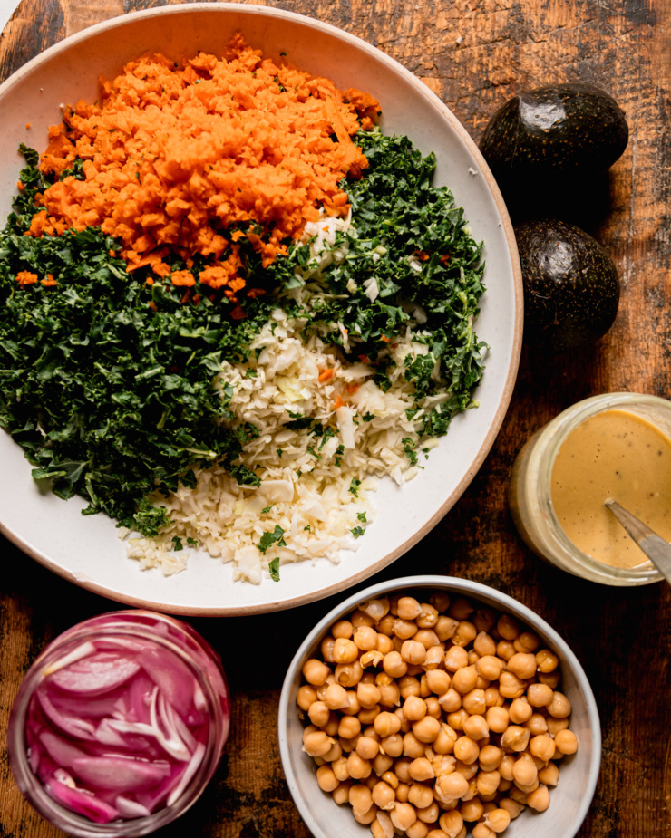 An overhead shot shows all prepped ingredients for a chopped kale cabbage salad: finely chopped kale, cabbage & carrots, ripe avocados, sweet mustard dressing, chickpeas, and pickled red onion.