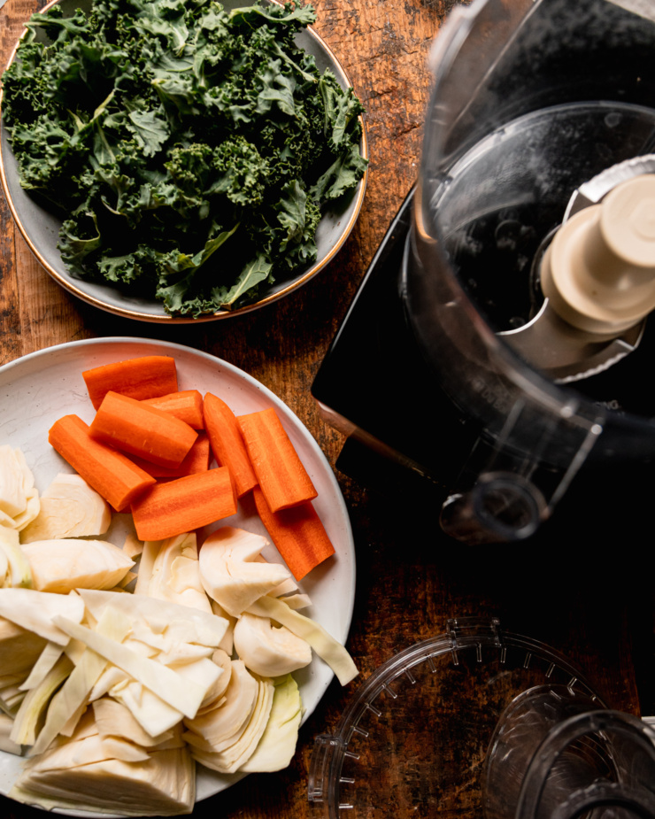 An overhead shot shows a food processor set up with kale leaves, chopped carrot, and roughly chopped cabbage nearby.