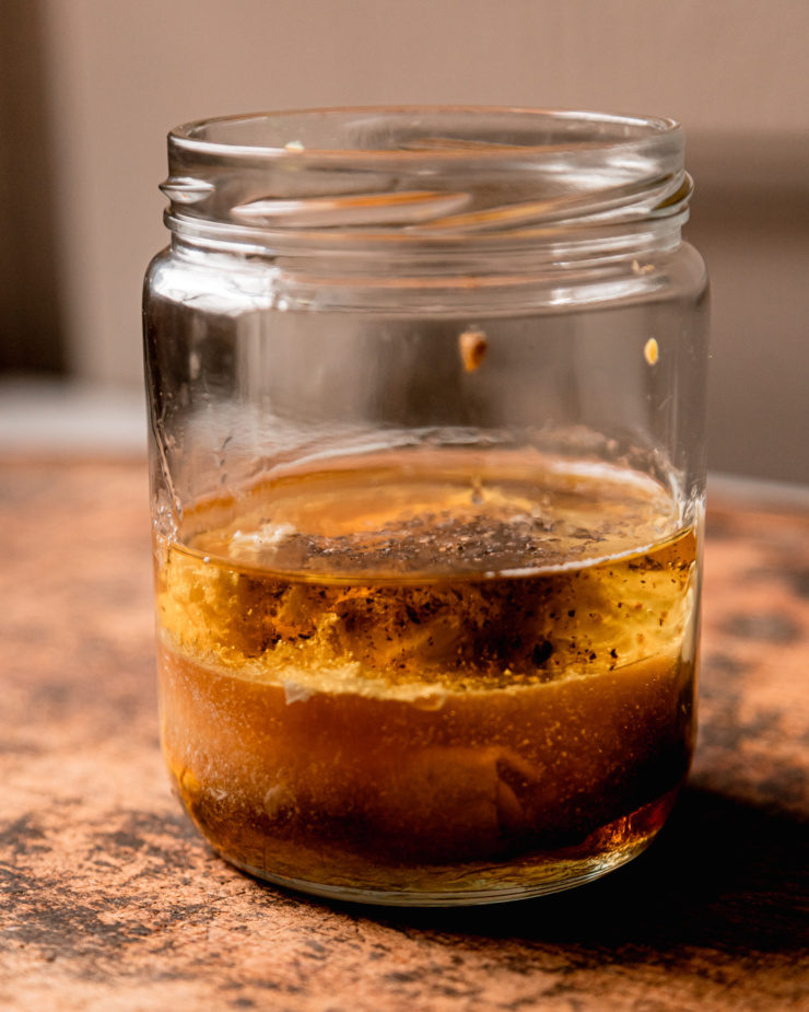 A head-on shot shows the layered ingredients for a sweet mustard dressing in a clear glass jar, prior to mixing.