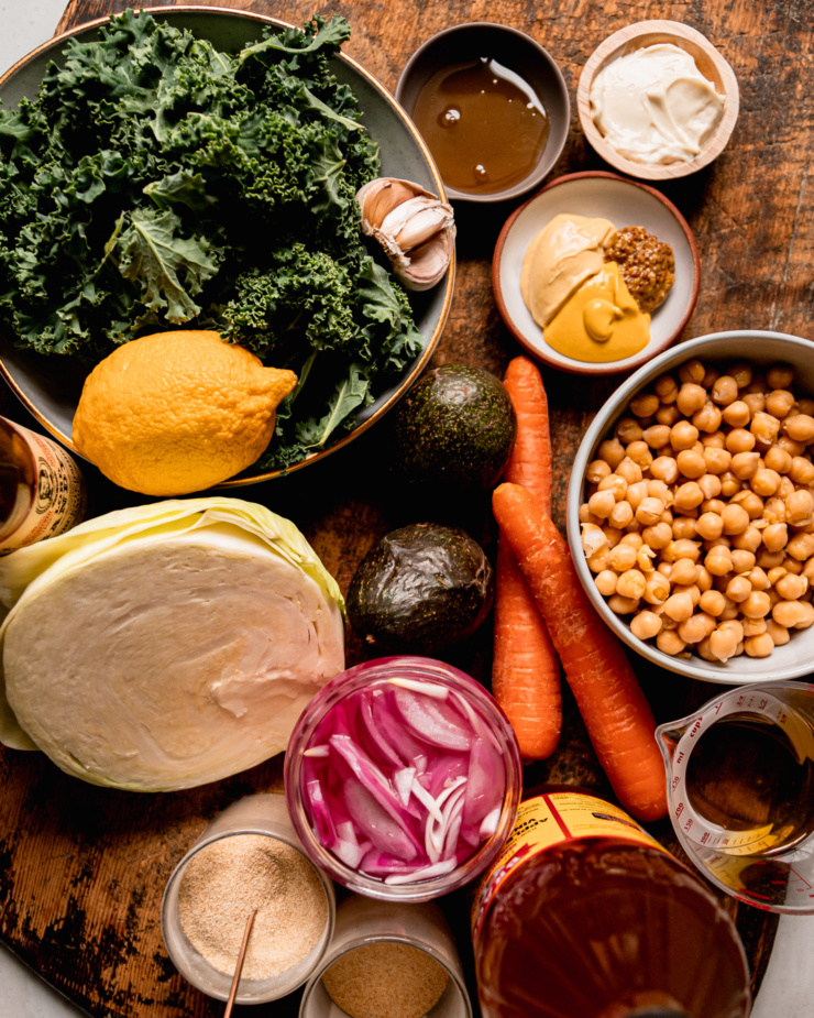An overhead shot shows ingredients for a chopped kale cabbage salad with sweet mustard dressing: maple syrup, vegan mayonnaise, 3 types of mustard, chickpeas, carrots, avocados, pickled red onions, avocado oil, apple cider vinegar, onion powder, garlic powder, cabbage, vegan Worcestershire sauce, lemon, and kale.