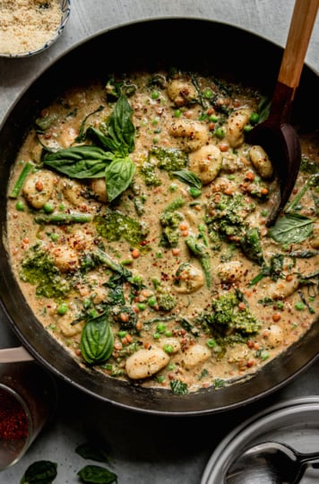 An overhead shot shows a creamy vegan gnocchi skillet with lentils, peas, and broccoli rabe. The dish is garnished with fresh basil and Aleppo chili flakes. A wooden serving spoon sticks out of the pot.