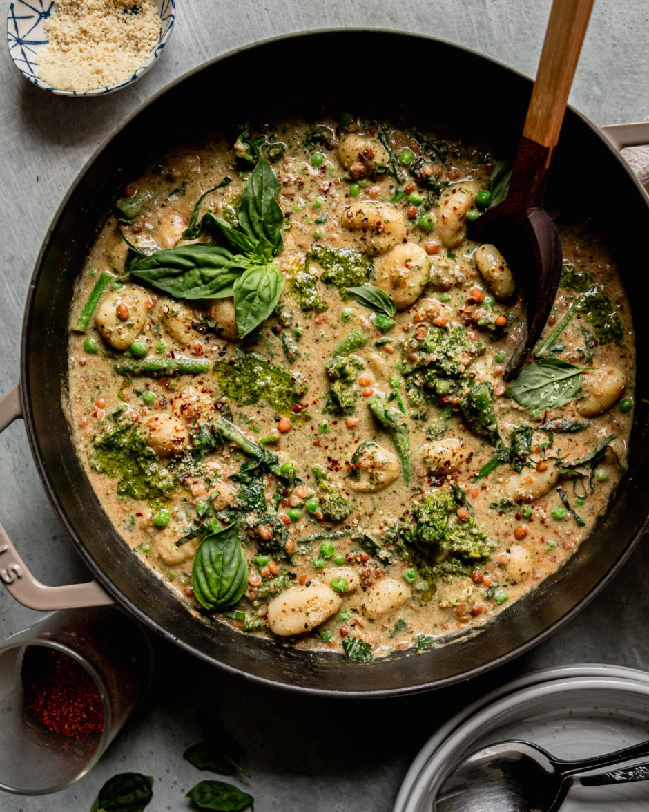 An overhead shot shows a creamy vegan gnocchi skillet with lentils, peas, and broccoli rabe. The dish is garnished with fresh basil and Aleppo chili flakes. A wooden serving spoon sticks out of the pot.