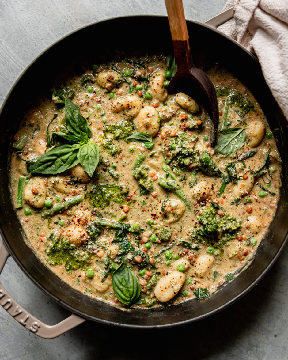 An overhead shot shows a creamy vegan gnocchi skillet with lentils, peas, and broccoli rabe. The dish is garnished with fresh basil and Aleppo chili flakes. A wooden serving spoon sticks out of the pot.