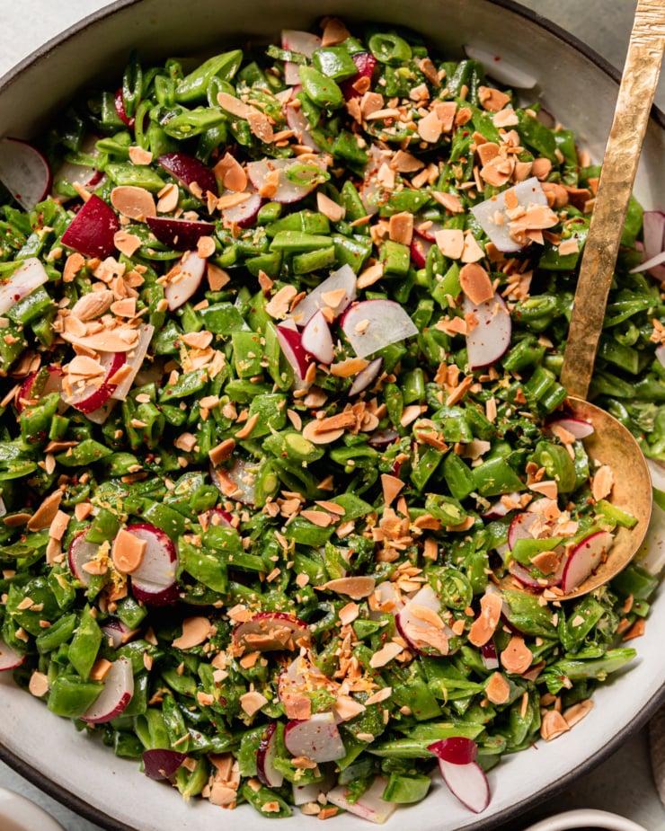 An up close, overhead shot shows a shredded snap pea salad with chopped radishes, mint, toasted almonds, and a lemon dressing. A brass ladle serving spoon is sticking out of the salad.