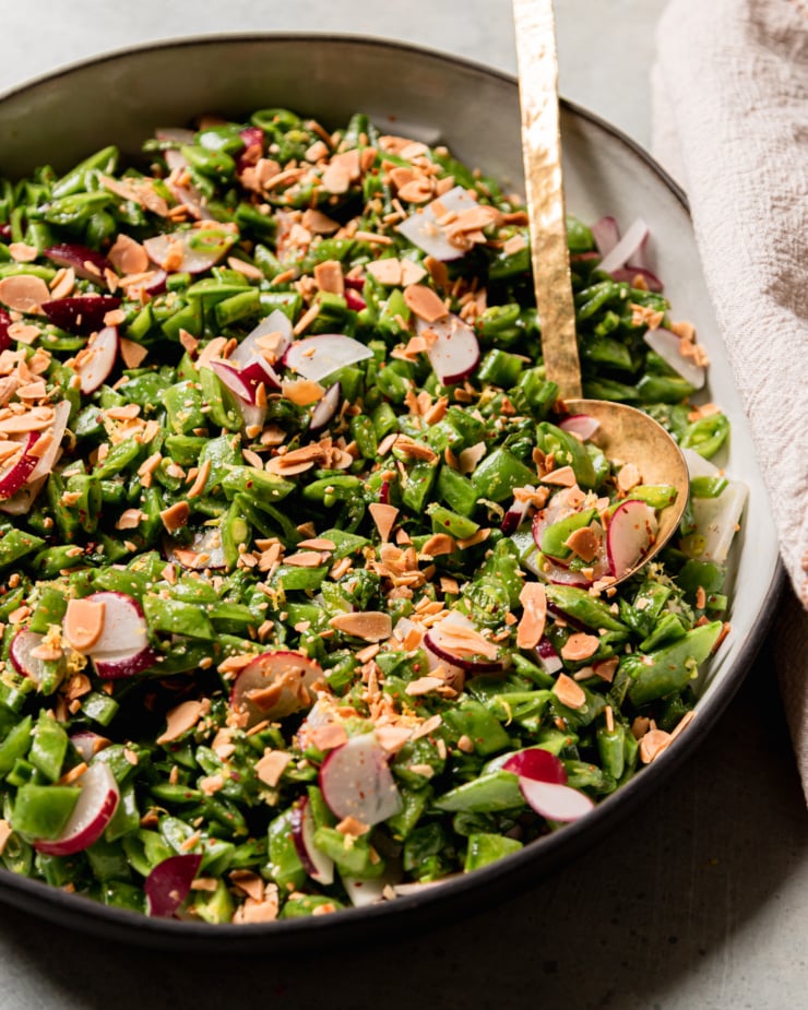 A 3/4 angle shot shows a shredded snap pea salad with chopped radishes, mint, toasted almonds, and a lemon dressing. A brass ladle serving spoon is sticking out of the salad.