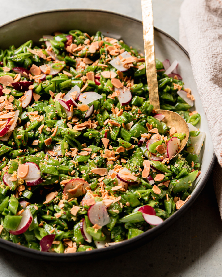 A 3/4 angle shot shows a shredded snap pea salad with chopped radishes, mint, toasted almonds, and a lemon dressing. A brass ladle serving spoon is sticking out of the salad.
