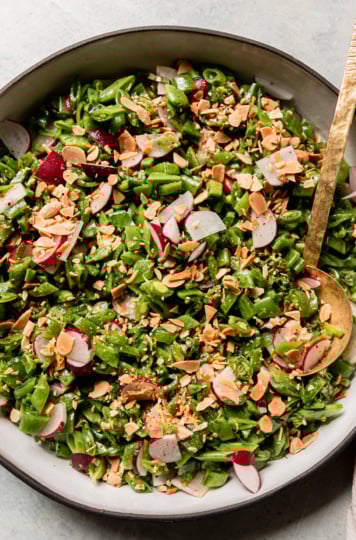 An overhead shot shows a shredded snap pea salad with chopped radishes, mint, toasted almonds, and a lemon dressing. A brass ladle serving spoon is sticking out of the salad.