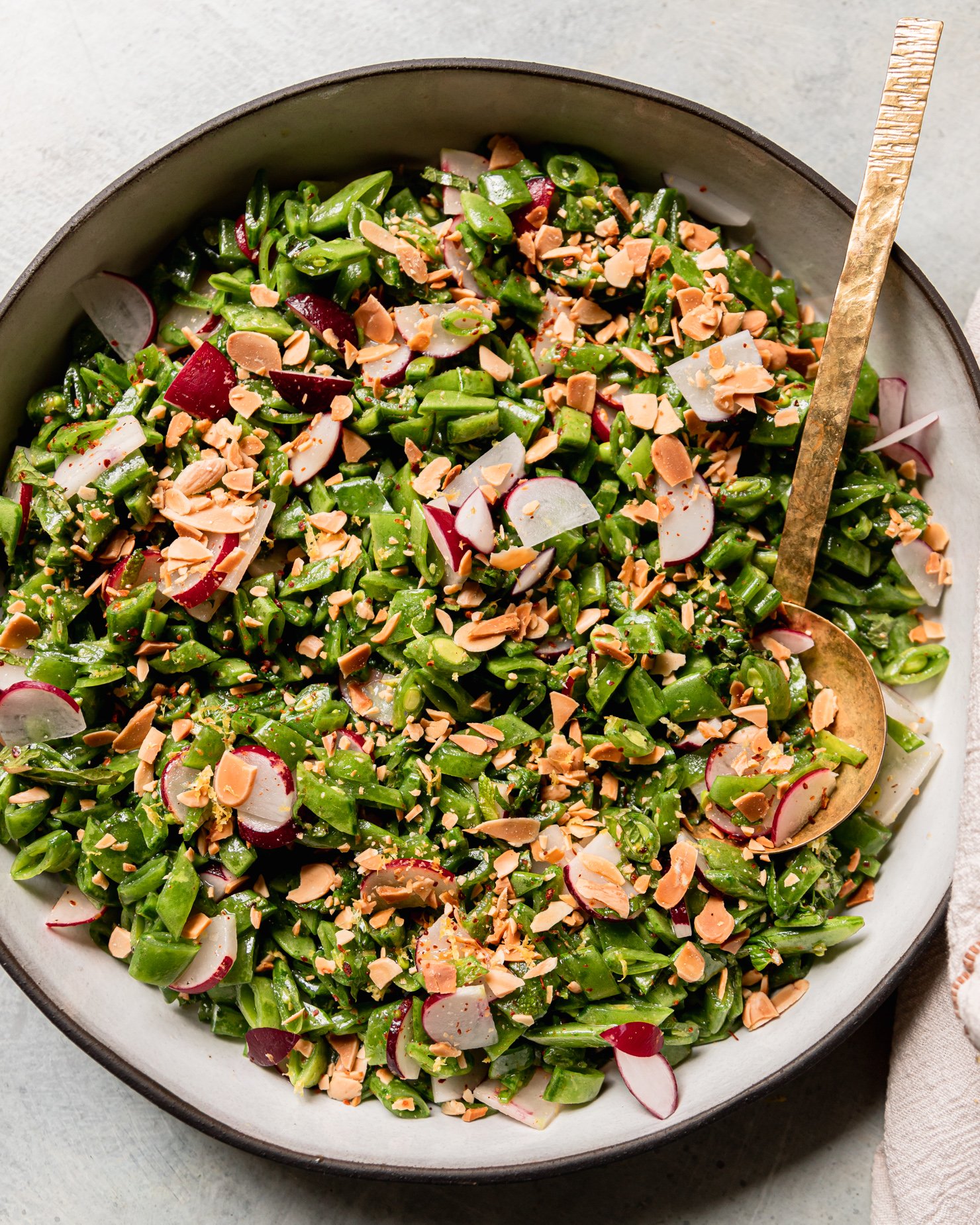 An overhead shot shows a shredded snap pea salad with chopped radishes, mint, toasted almonds, and a lemon dressing. A brass ladle serving spoon is sticking out of the salad.