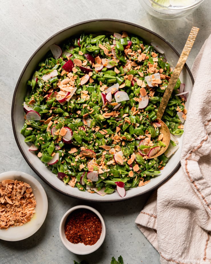 An up close, overhead shot shows a shredded snap pea salad with chopped radishes, mint, toasted almonds, and a lemon dressing. A brass ladle serving spoon is sticking out of the salad. Bowls of aleppo pepper and chopped toasted almonds are nearby.
