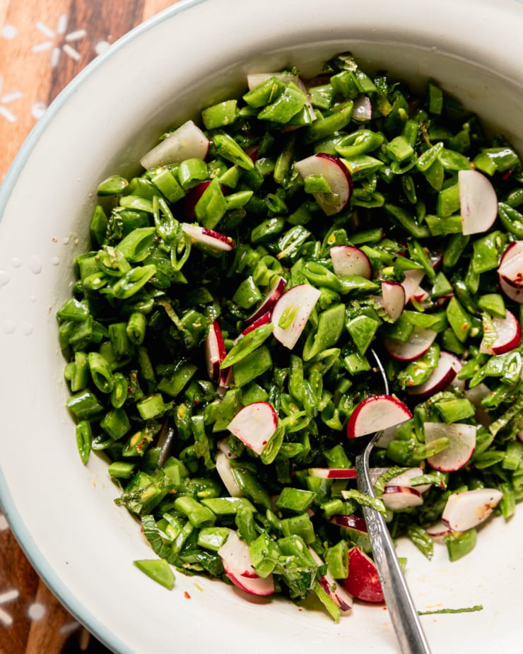 An overhead shot shows a salad of finely chopped snap peas, radishes, mint, and lemony dressing. The salad is in an enamelware bowl.