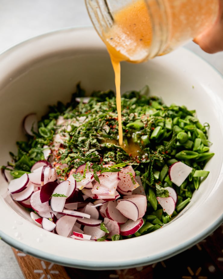A 3/4 angle shot shows salad dressing being poured over a mixture of chopped radishes, snap peas, chopped mint, and a sprinkle of Aleppo pepper.