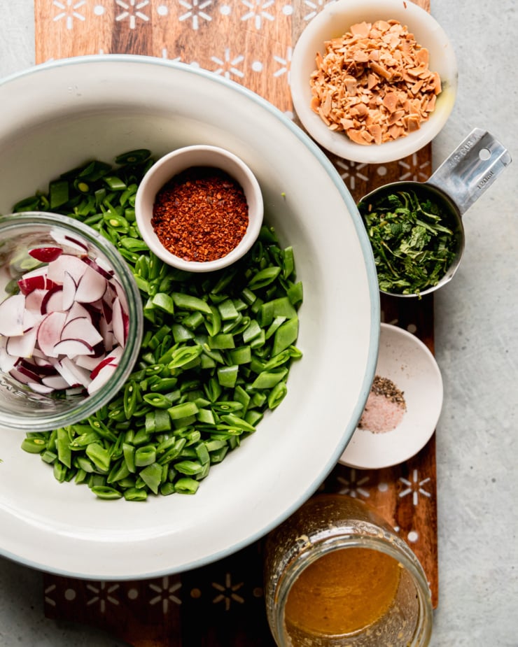AN overhead shot shows prepped ingredients for a salad: toasted chopped almonds, chopped mint, salt and pepper, lemony miso dressing, finely chopped snap peas, chopped radishes, and Aleppo pepper.