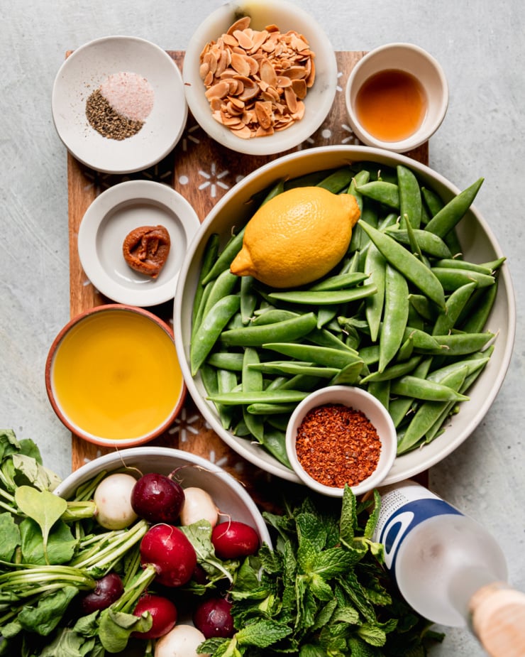 An overhead shot shows ingredients for a snap pea salad: maple syrup, snap peas, lemon, Aleppo pepper, champagne vinegar, fresh mint, radishes, olive oil, miso, salt, pepper, and toasted chopped almonds.