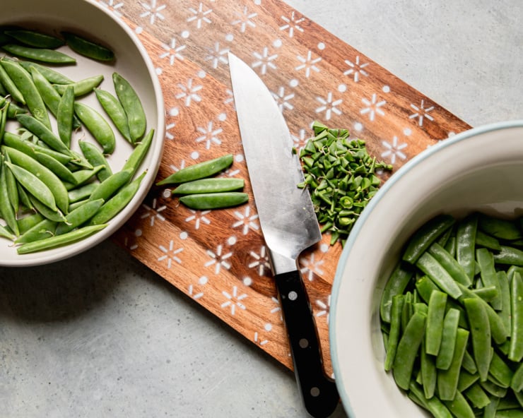 An overhead shot shows the process of snap pea ends being removed on a cutting board.