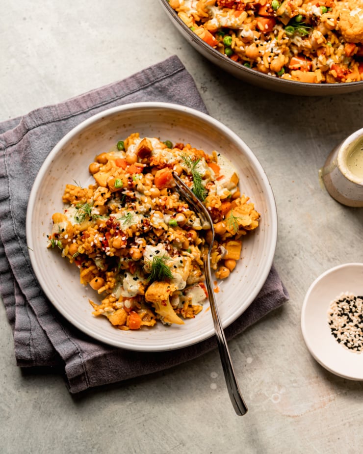 An overhead shot shows a serving of rice and veggies with chickpeas and a dilly tahini sauce. The serving is garnished with fresh dill, chili crisp and sesame seeds.