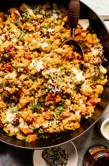 An overhead shot shows one-pot rice and veggies with chickpeas and a drizzle of creamy dilly tahini sauce on top. The dish is garnished with fresh dill, chili crisp and sesame seeds. A serving spoon is sticking out of the pot.