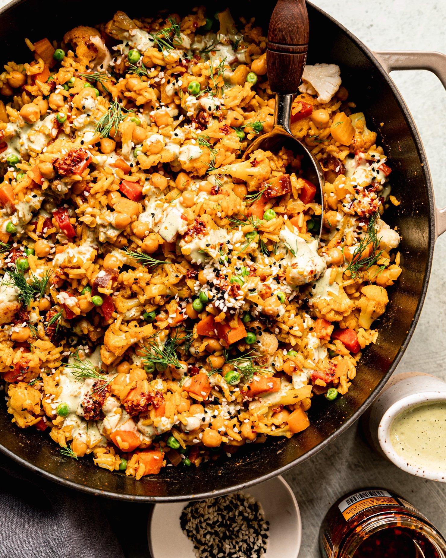 An overhead shot shows one-pot rice and veggies with chickpeas and a drizzle of creamy dilly tahini sauce on top. The dish is garnished with fresh dill, chili crisp and sesame seeds. A serving spoon is sticking out of the pot.