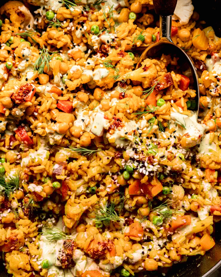 An up close, overhead shot shows one-pot rice and veggies with chickpeas and a drizzle of creamy dilly tahini sauce on top. The dish is garnished with fresh dill, chili crisp and sesame seeds. A serving spoon is sticking out of the pot.