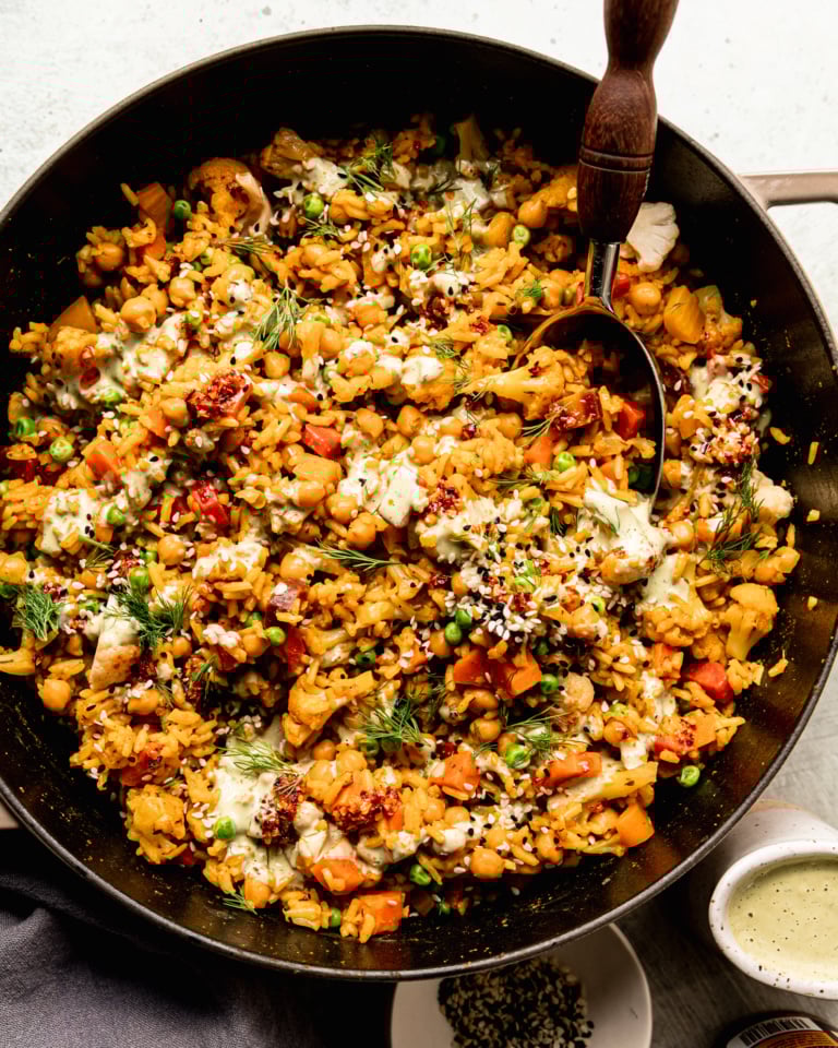 An overhead shot shows one-pot rice and veggies with chickpeas and a drizzle of creamy dilly tahini sauce on top. The dish is garnished with fresh dill, chili crisp and sesame seeds. A serving spoon is sticking out of the pot.