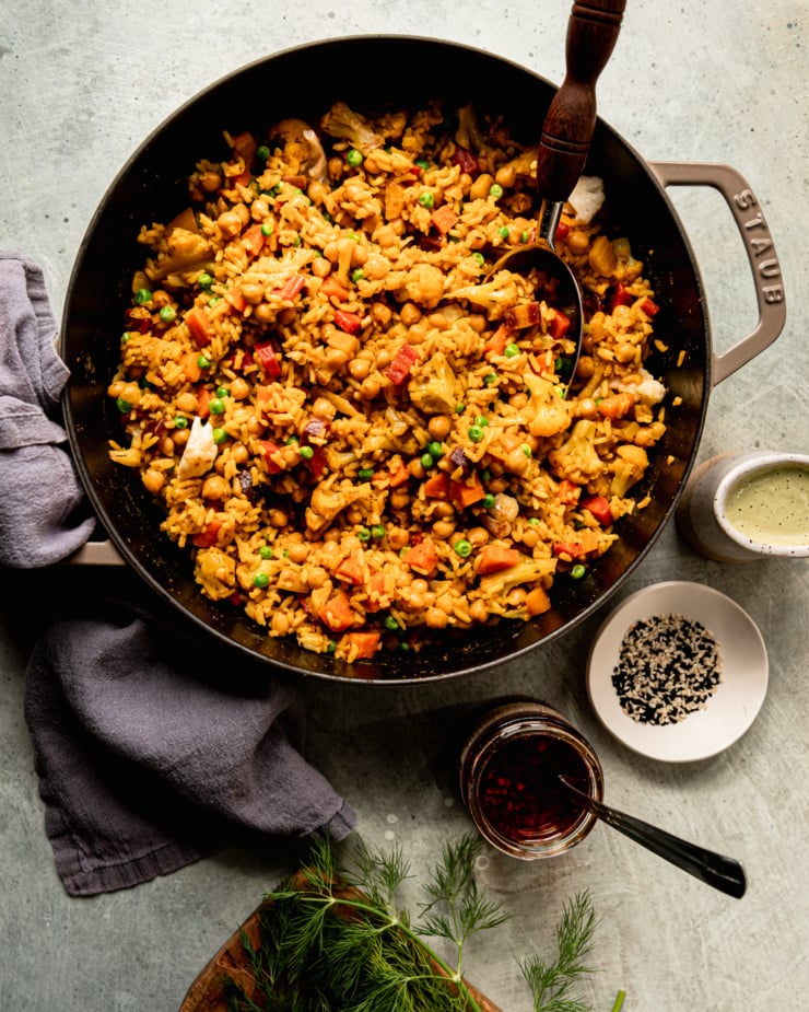 An overhead shot shows one-pot rice and veggies with chickpeas. A jar of chili crisp, a bowl of sesame seeds, and a cruet of dilly tahini sauce are all nearby. A serving spoon is sticking out of the pot and a linen napkin is nearby.