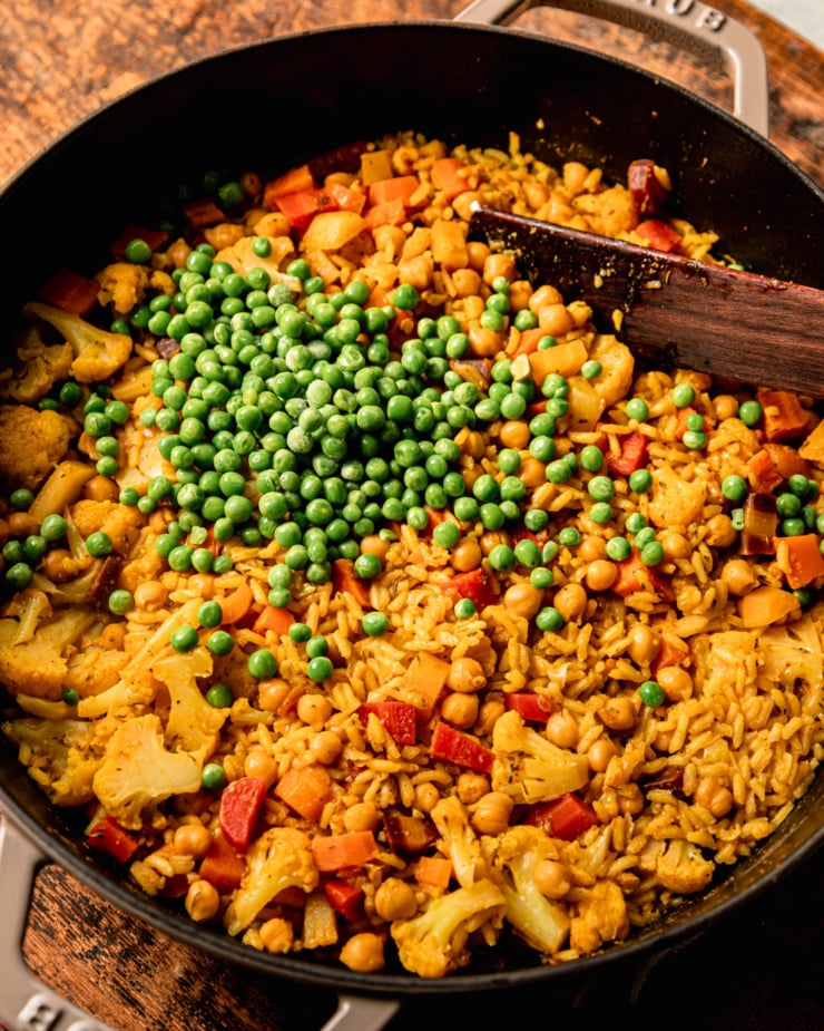 An overhead shot shows a pot of rice and veggies with chickpeas and frozen peas poured on top.