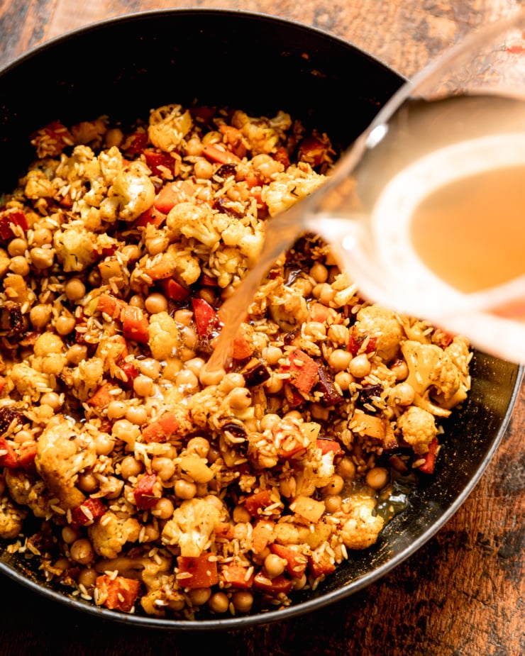 An overhead shot shows vegetable stock being poured into a pot of vegetables, rice, and chickpeas.