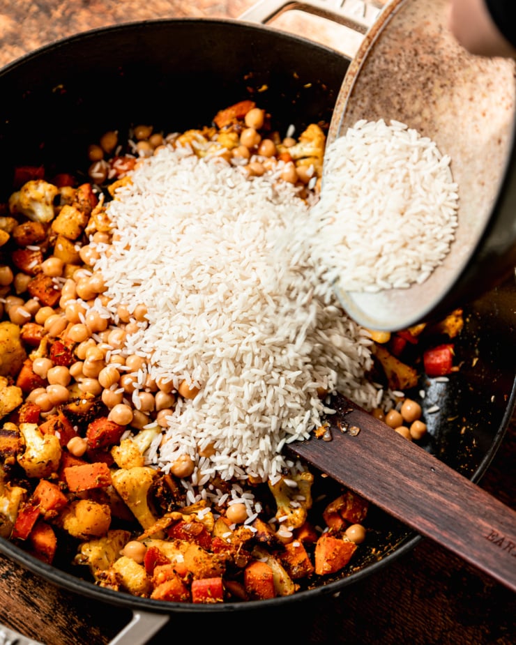 An overhead shot shows jasmine rice being poured into a pot with sautรฉed carrots and cauliflower and chickpeas.