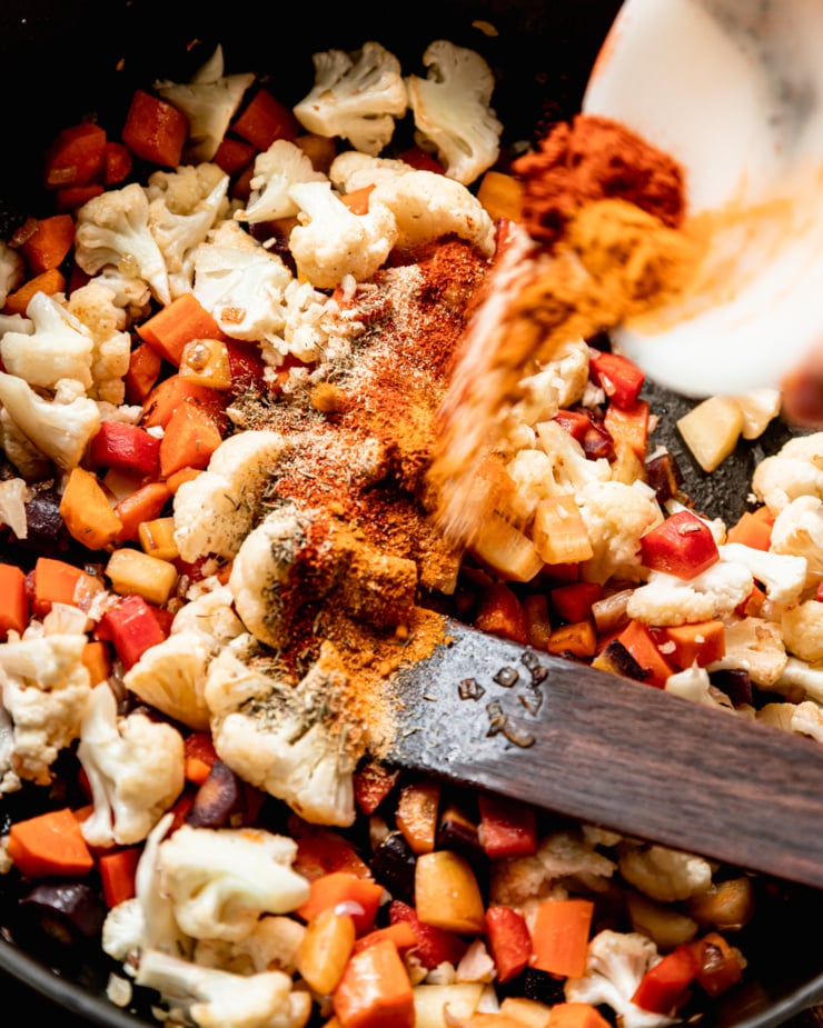 An overhead shot shows spices being poured into a pot with sautรฉed cauliflower and carrots.