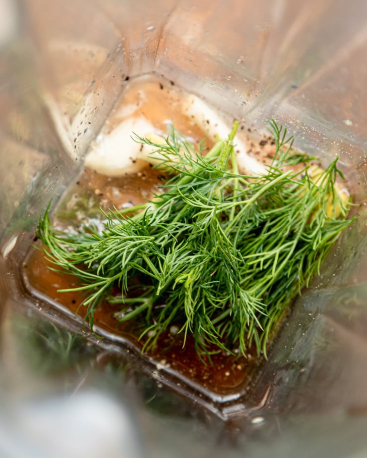 An overhead shot shows dill, garlic, tahini, water, and lemon juice in a blender pitcher.
