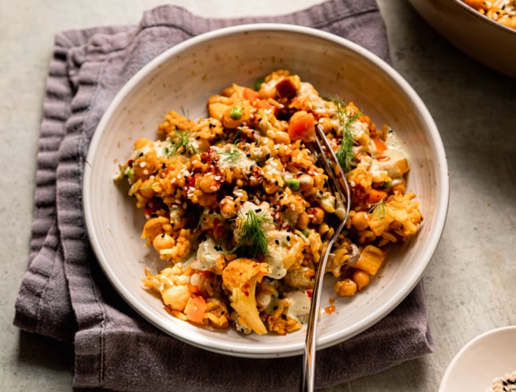 An overhead shot shows a serving of rice and veggies with chickpeas and a dilly tahini sauce. The serving is garnished with fresh dill, chili crisp and sesame seeds.