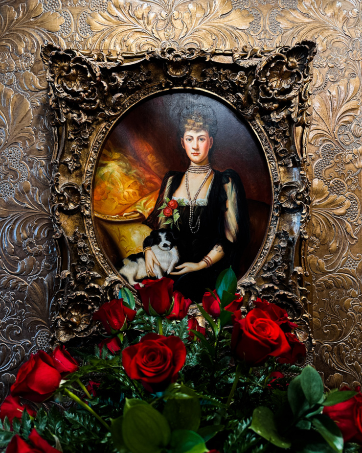 A head-on shot shows a framed portrait of a woman and her small dog. A bouquet of roses is in front of the portrait hanging on the wall.