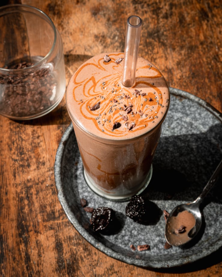 An overhead shot shows a glass of vegan chocolate fantasy smoothie with almond butter swirls and cacao nibs on top. Dried prunes are seen nearby. A straw is sticking out of the smoothie.
