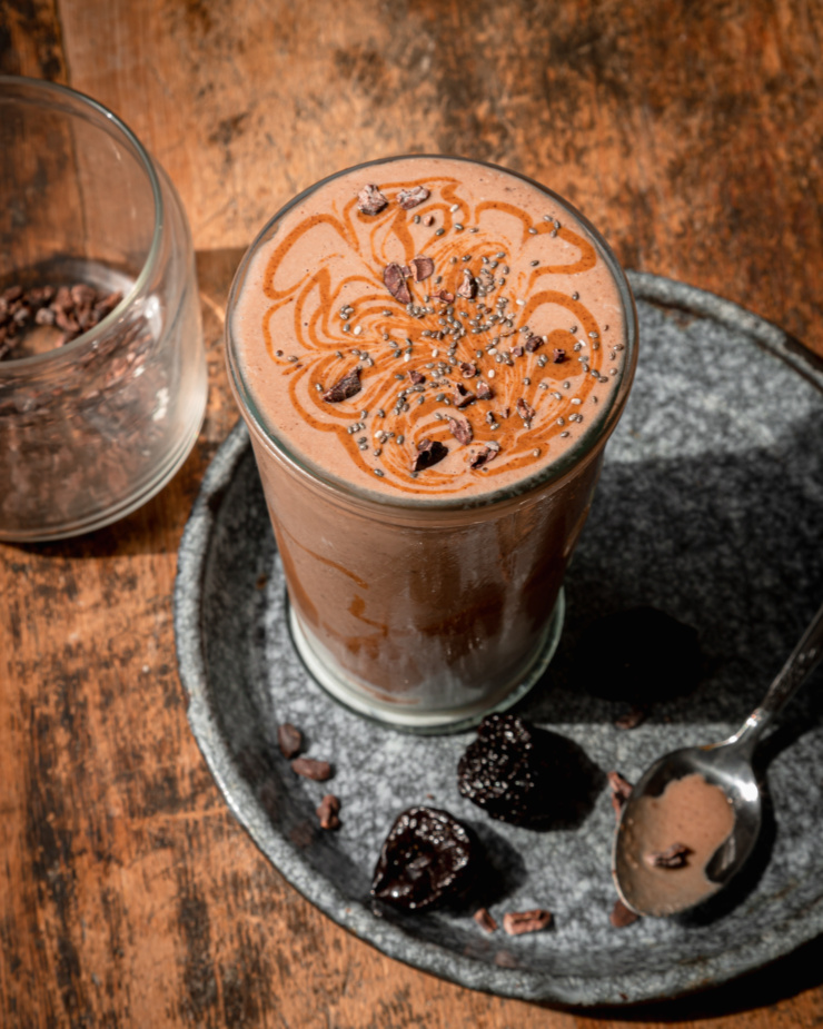 An overhead shot shows a glass of chocolate fantasy smoothie with almond butter swirls and cacao nibs on top. Dried prunes are seen nearby.