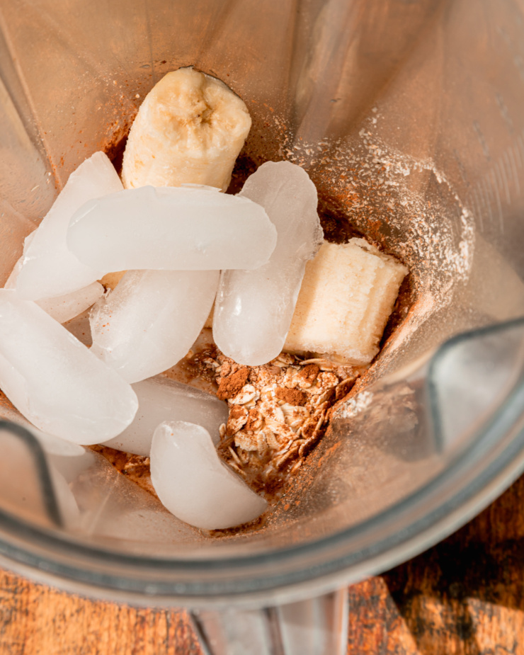 An overhead shot shows ice, frozen banana, oats, cocoa powder and other smoothie ingredients in a blender pitcher.