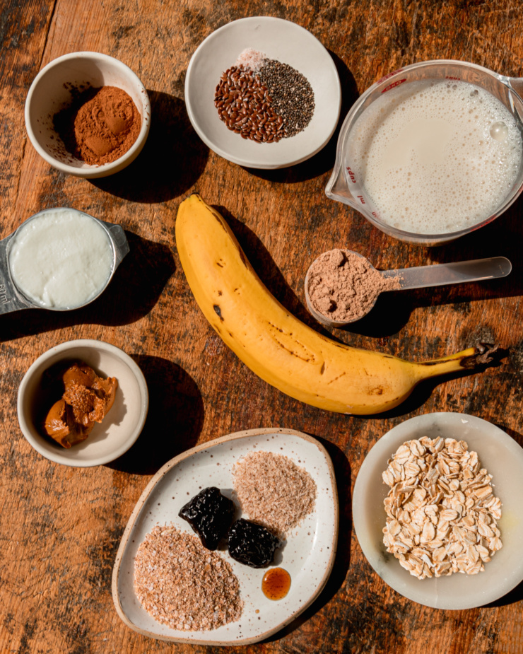 An overhead shot shows ingredients for a vegan chocolate smoothie recipe: chia seeds, flax seeds. salt, almond milk, chocolate protein powder, banana, oats, psyllium husk, prunes, wheat bran, vanilla, almond butter, vegan yogurt, and cocoa powder.