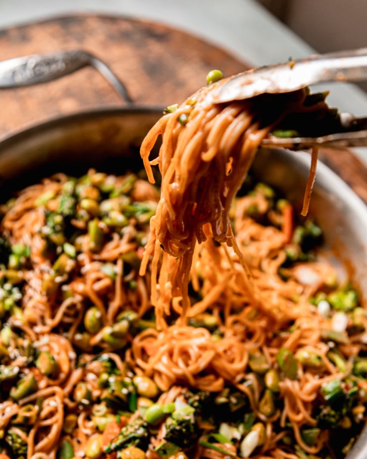 An overhead shot shows a apir of tongs lifting some Gochujang noodles out of a skillet.