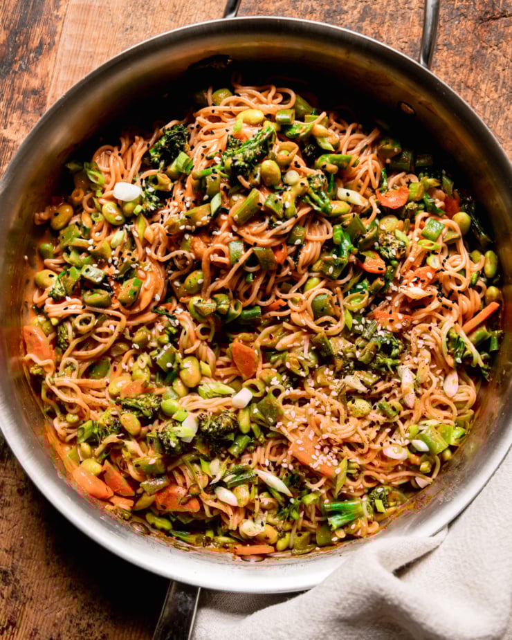 An overhead shot shows a bunch of vegan Gochujang noodles in a skillet with lots of sautéed chopped vegetables. The noodles are garnished with sliced green onions and sesame seeds.