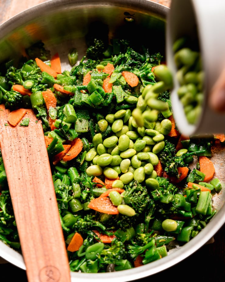 An overhead shot shows thawed and shelled edamame being poured into a pan with sautéed chopped vegetables.
