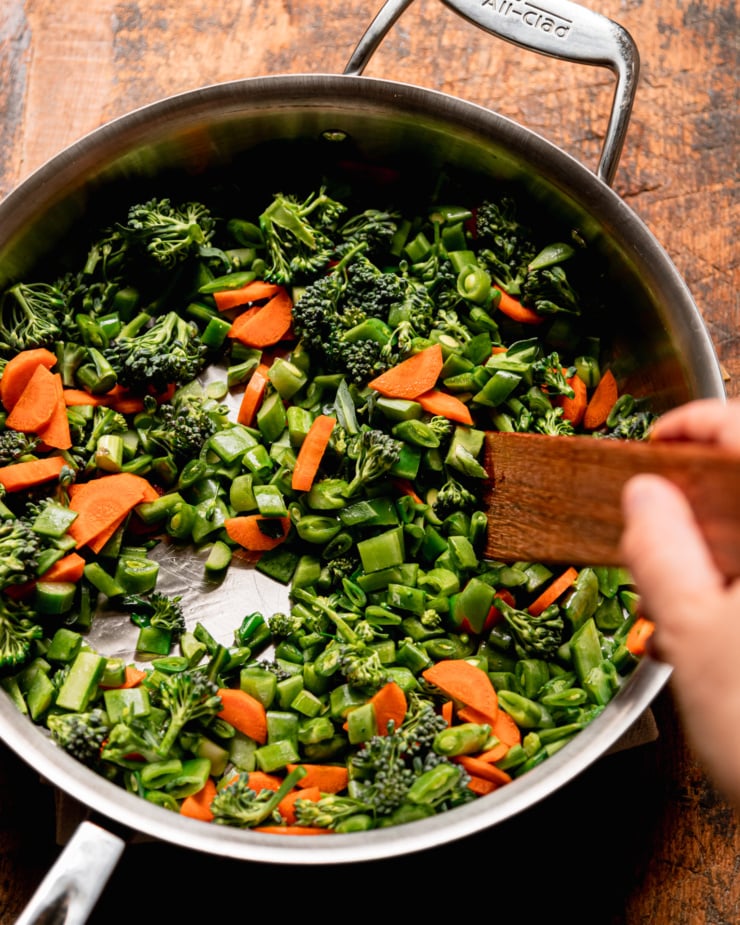 An overhead shot shows a hand using a wooden utensil to stir up fine cuts of vegetables in a sauté pan.