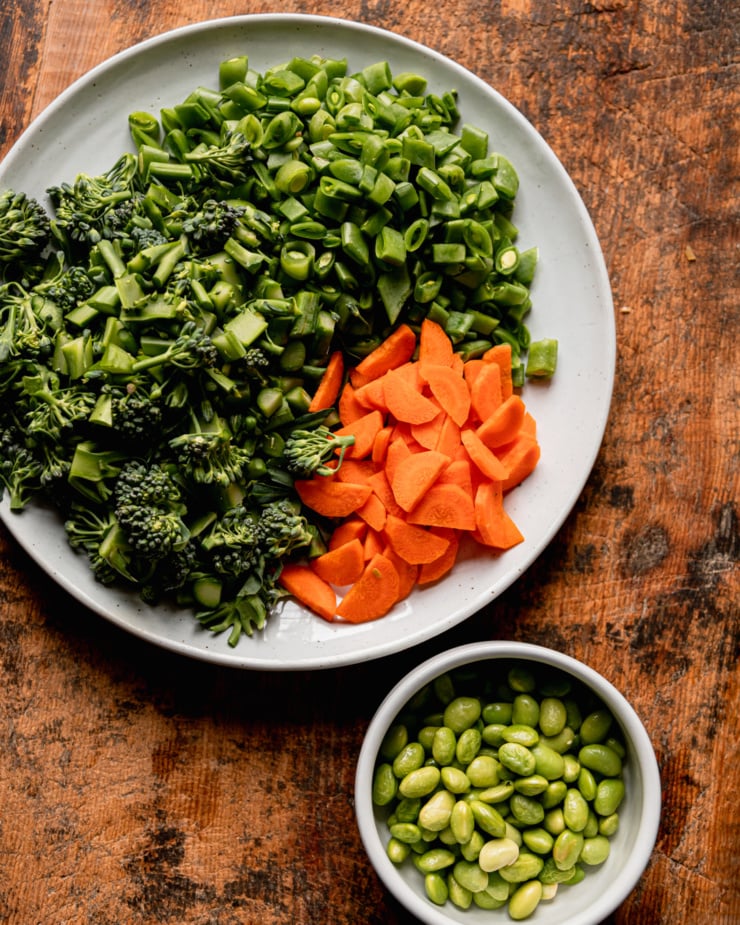 An overhead shot shows a plate with chopped snap peas, broccolini, and carrots. A bowl of thawed frozen edamame is nearby.
